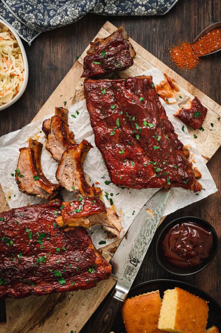 Oven baked baby back ribs in the process of being cut into individual ribs on a cutting board with butcher paper and barbecue sauce.