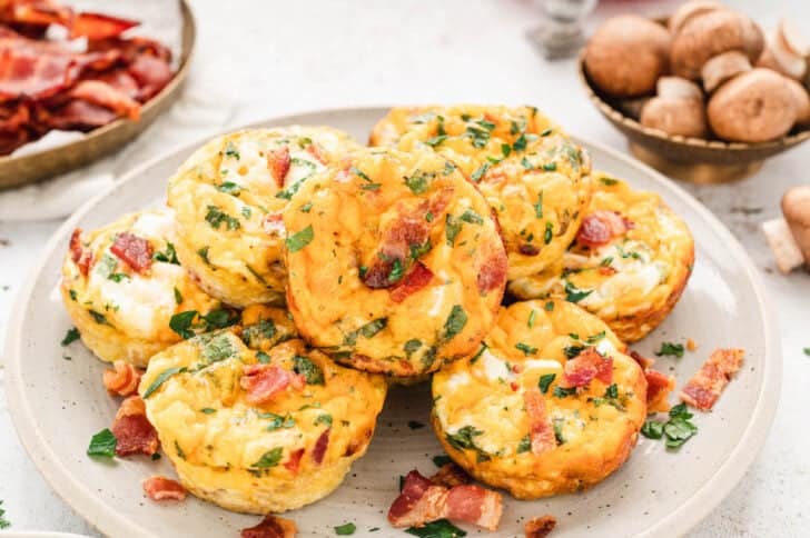 Egg muffins made with bacon, cheese and veggies piled up on a white plate.