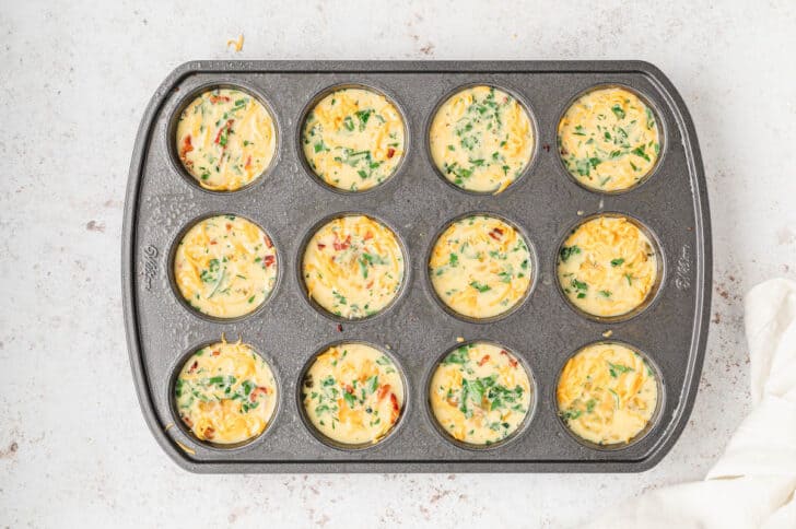 A muffin pan with the holes filled with raw breakfast egg muffins.
