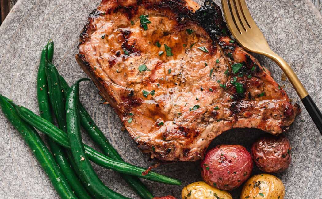 A grilled pork chop on a textured gray plate alongside roasted baby potatoes and green beans.