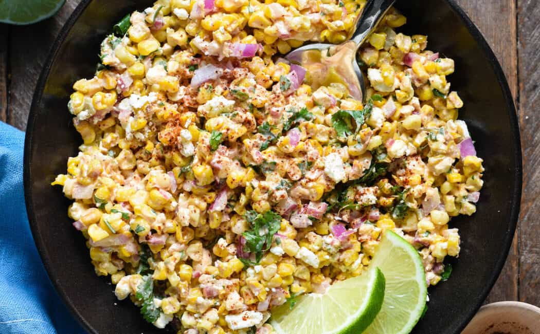 Mexican roasted corn salad in a black bowl, garnished with lime wedges.