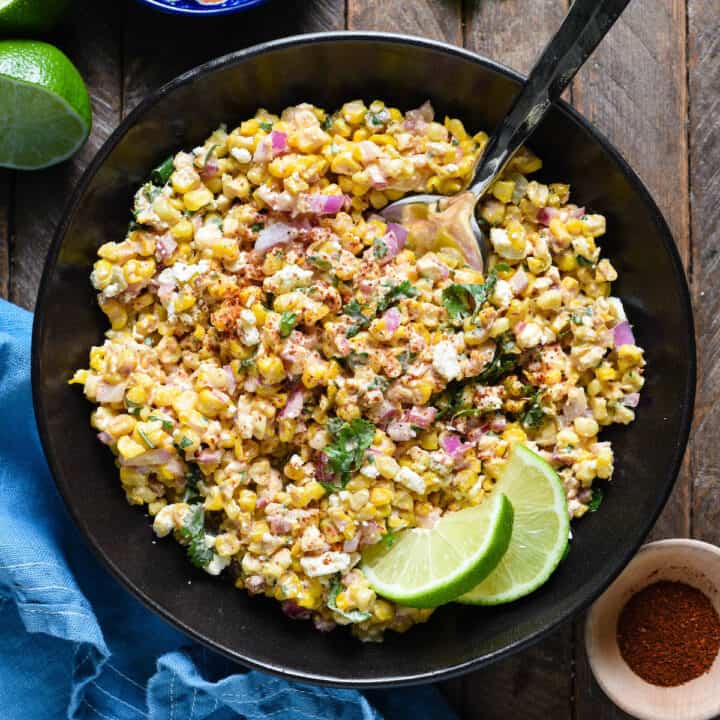 Mexican roasted corn salad in a black bowl, garnished with lime wedges.