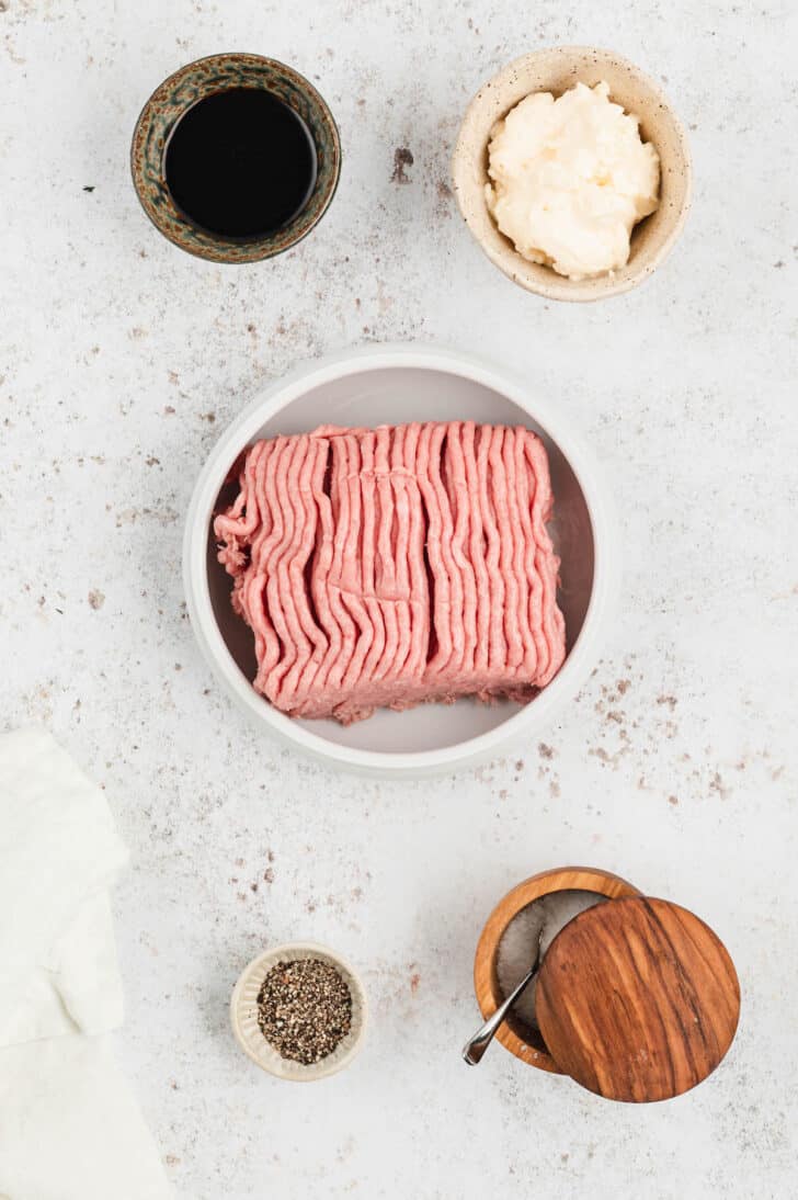 Ingredients laid out in bowls on a light surface, including ground poultry, Worcestershire sauce, mayonnaise, and salt and pepper.