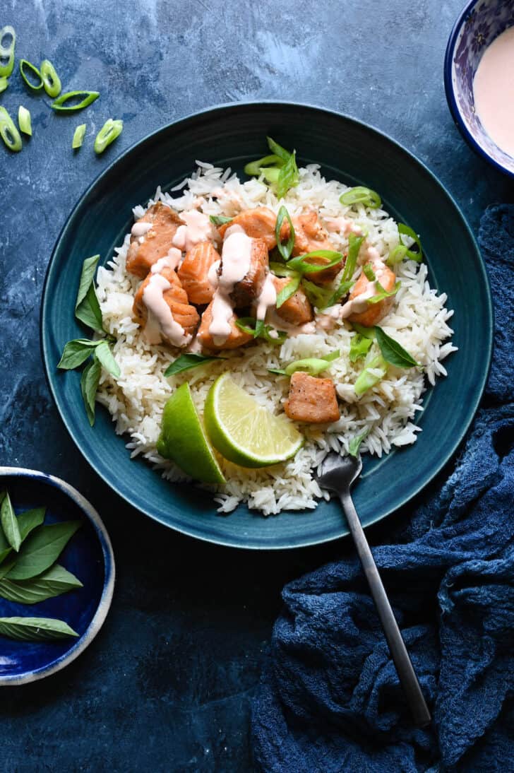 A blue bowl filled with white rice, air fryer salmon bites topped with a creamy sauce, sliced green onions and lime wedges, with a fork sticking into the rice.