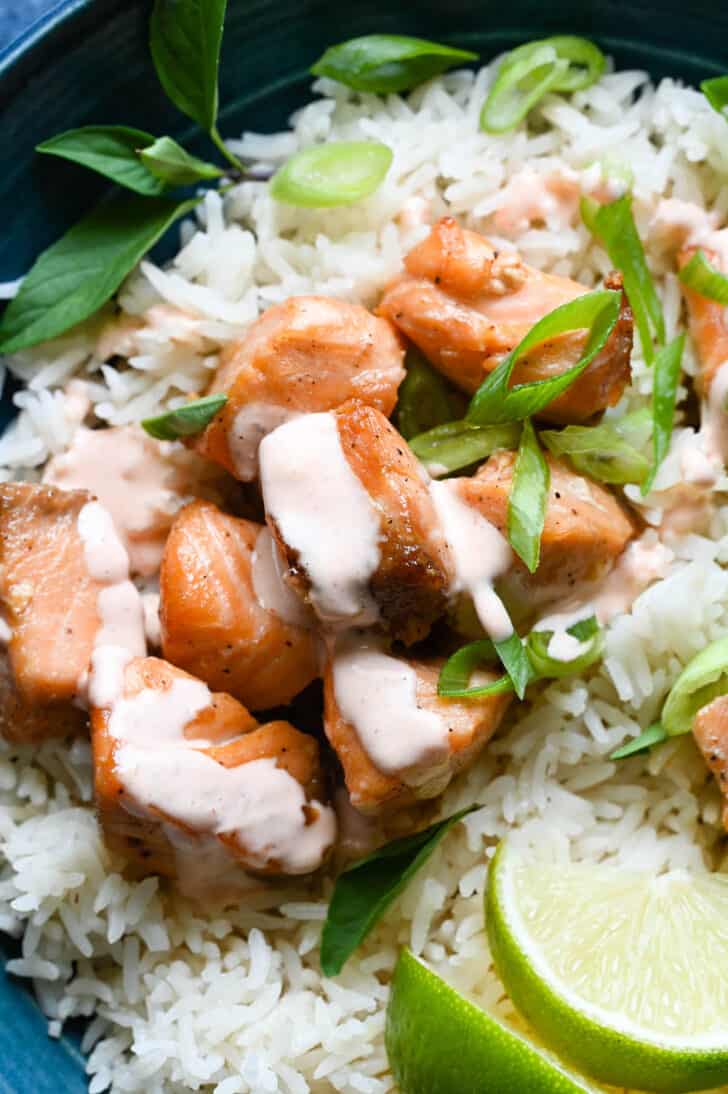 A blue bowl filled with white rice, air fryer salmon bites topped with a creamy sauce, sliced green onions and lime wedges.