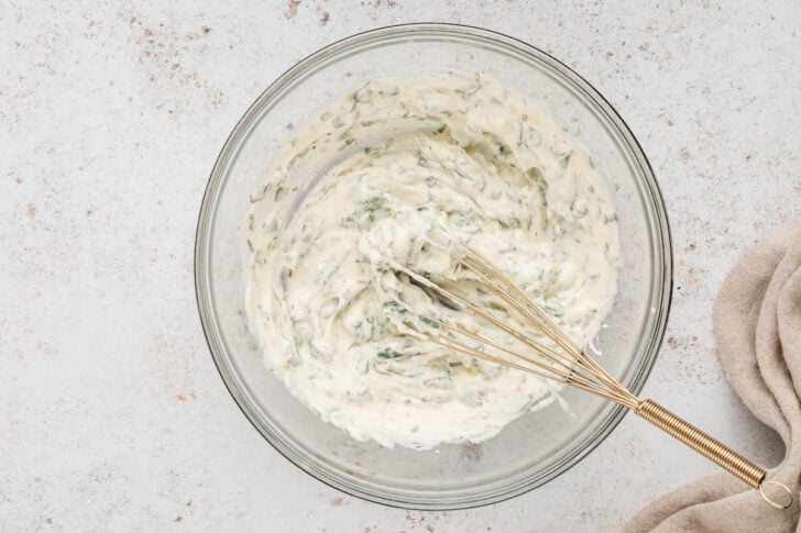 A glass bowl filled with cilantro mayonnaise with a whisk in it.