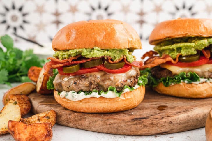 Two avocado burgers on a brioche bun stopped with bacon, lettuce, tomato and pickled jalapeno. The burgers are on wooden cutting board in front of a patterned background. There are potato wedges in the foreground.