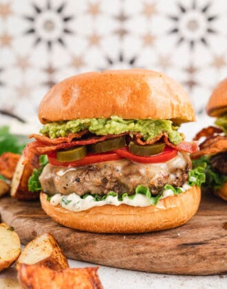 A bacon avocado burger on a brioche bun topped with lettuce, tomato and pickled jalapeno. The burger is on wooden cutting board in front of a patterned background. There are potato wedges in the foreground.