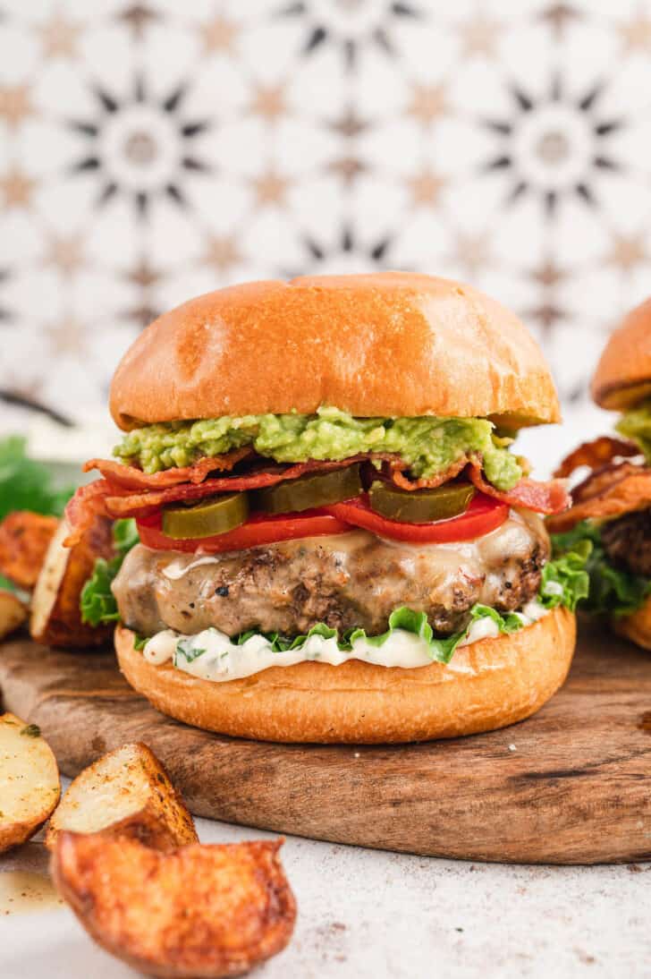 A bacon avocado burger on a brioche bun topped with lettuce, tomato and pickled jalapeno. The burger is on wooden cutting board in front of a patterned background. There are potato wedges in the foreground.