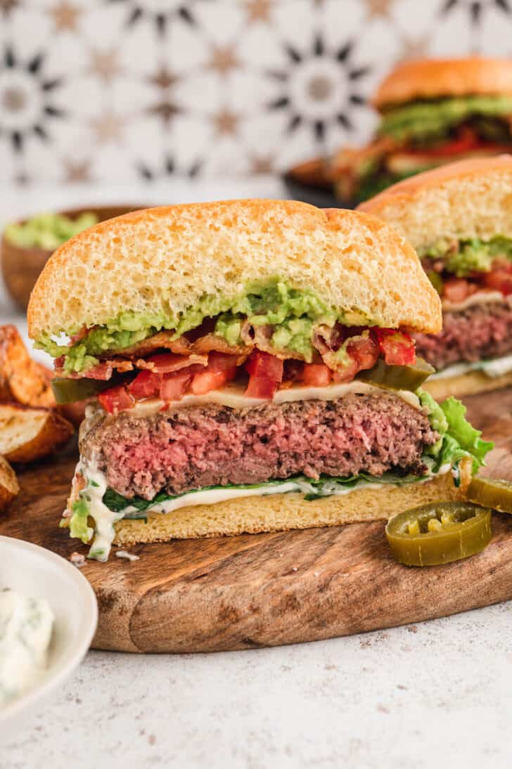 A cross-section of a cut-in-half cheeseburger topped with guacamole, bacon, lettuce, tomato and cilantro mayo.