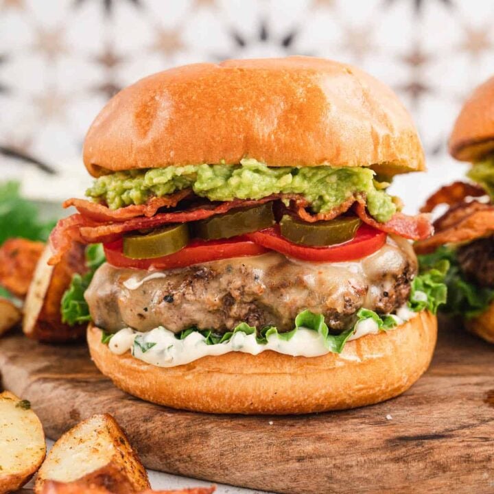 A bacon avocado burger on a brioche bun topped with lettuce, tomato and pickled jalapeno. The burger is on wooden cutting board in front of a patterned background. There are potato wedges in the foreground.