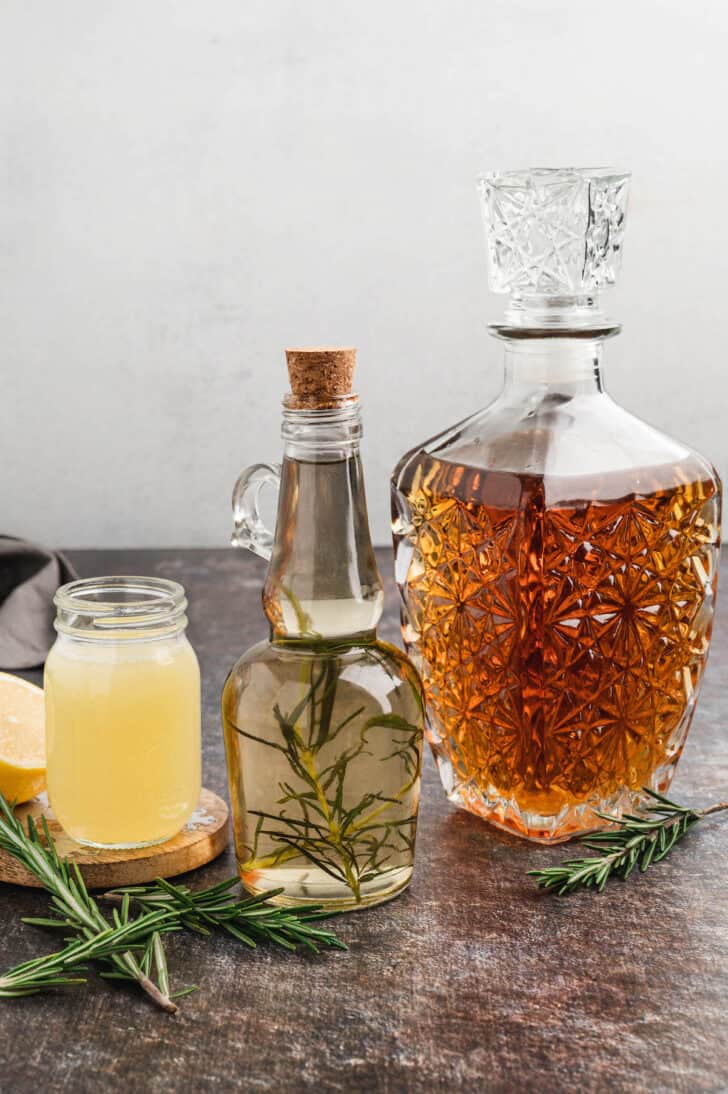 Ingredients needed for a bourbon sour recipe, including whiskey in a fancy glass bottle, rosemary simple syrup and squeezed lemon juice.
