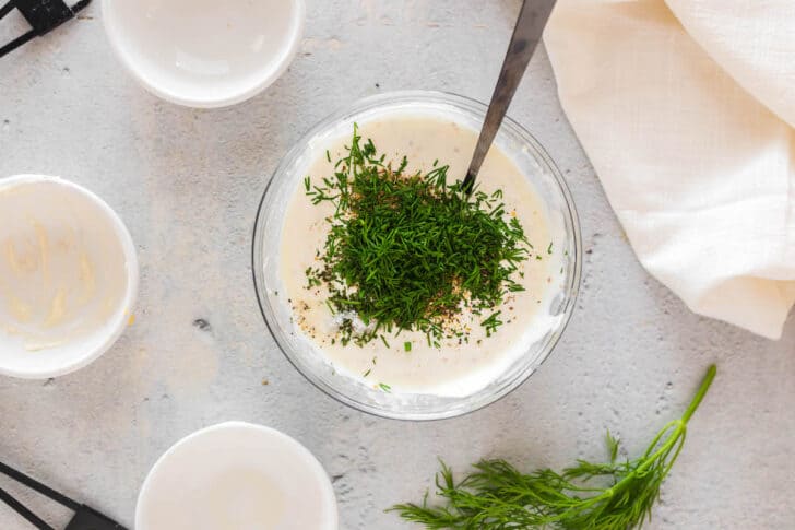 A glass bowl filled with a white sauce, topped with chopped dill.