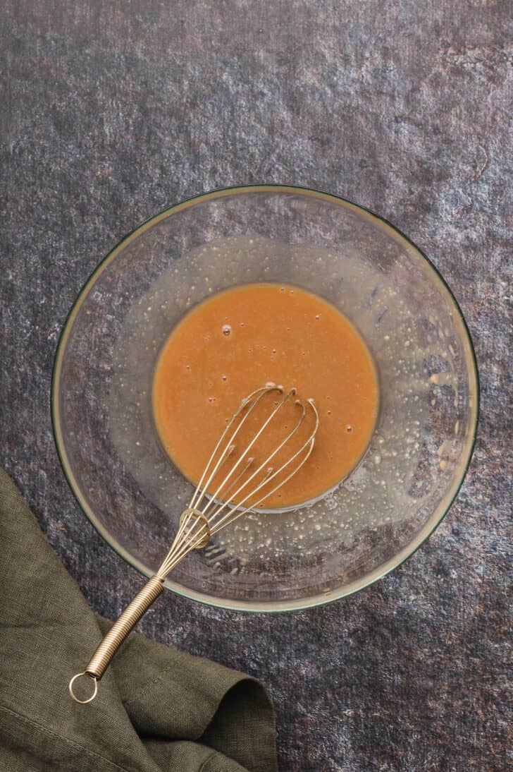 A glass bowl with a whisk stirring together a light brown dressing.
