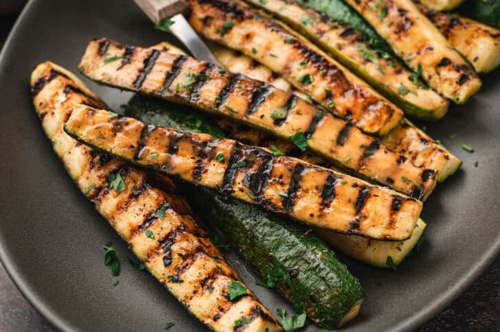 Grilled zucchini spears with a miso glaze on a dark gray oval platter with a serving fork.