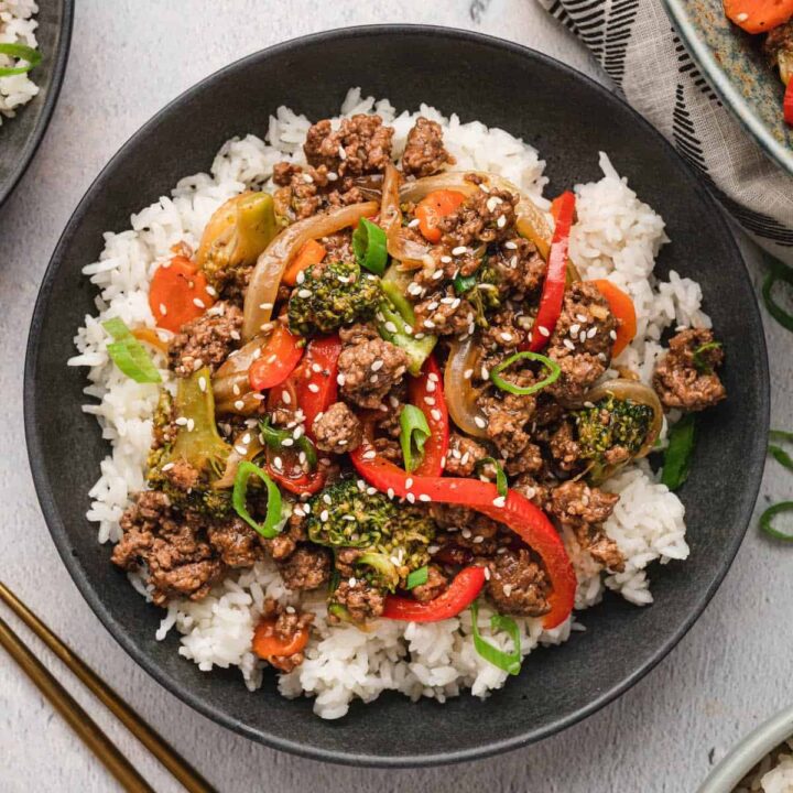 A black bowl filled with white rice topped with ground beef stir fry and veggies, garnished with green onions and sesame seeds.