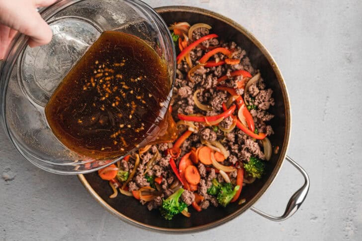 A hand pouring a brown liquid into a skillet filled with meat and vegetables.