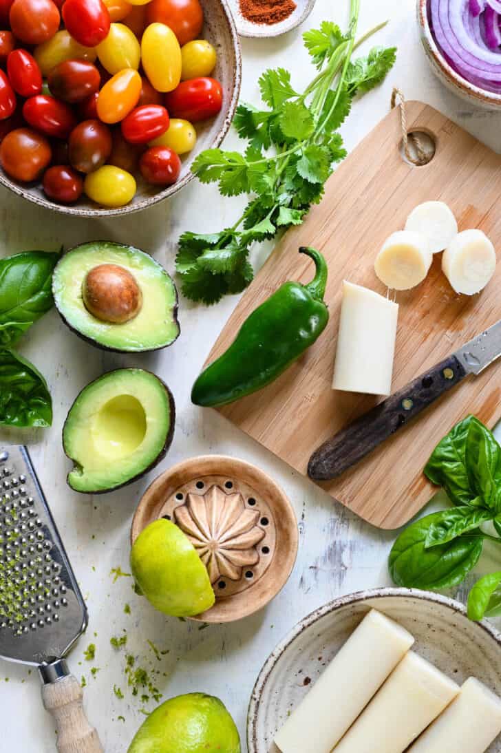 Ingredients on a light surface including tomatoes, jalapeno, avocado, herbs and palmito.