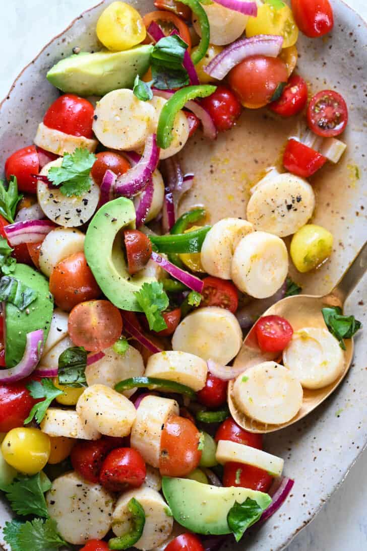 A speckled oval platter filled with heart of palm salad with tomatoes and avocado, with a gold spoon scooping some out.