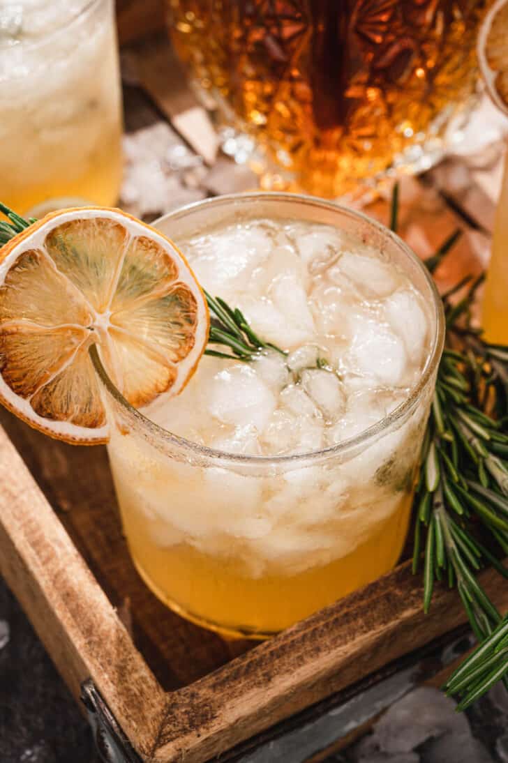 Closeup on a whiskey drink over crushed ice, garnished with an herb sprig and dried citrus slice.