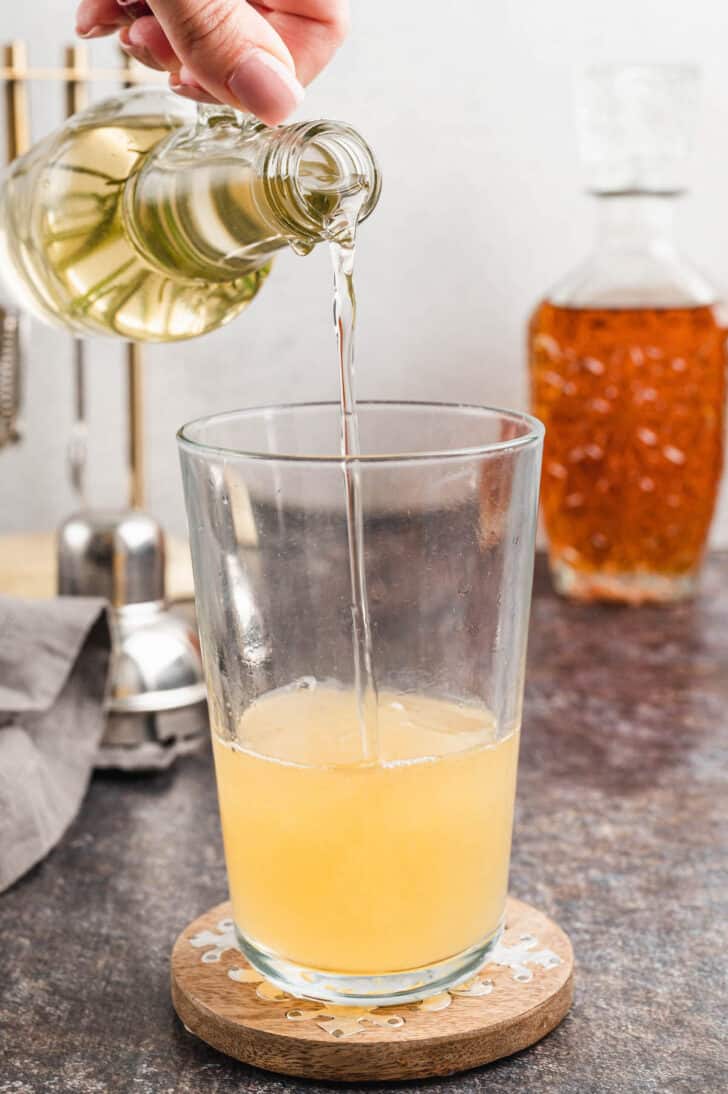 A hand pouring a light colored syrup into a tall glass filled with light orange juice.