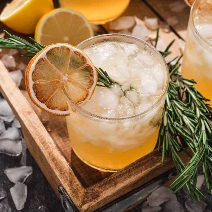 A rosemary cocktail filled with crushed ice, a rosemary sprig and a dried lemon wheel, on a wooden tray.
