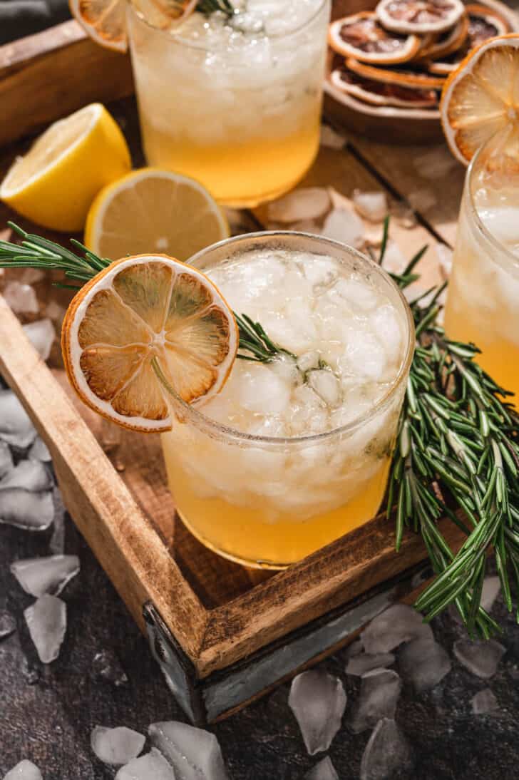 A rosemary cocktail filled with crushed ice, a rosemary sprig and a dried lemon wheel, on a wooden tray.