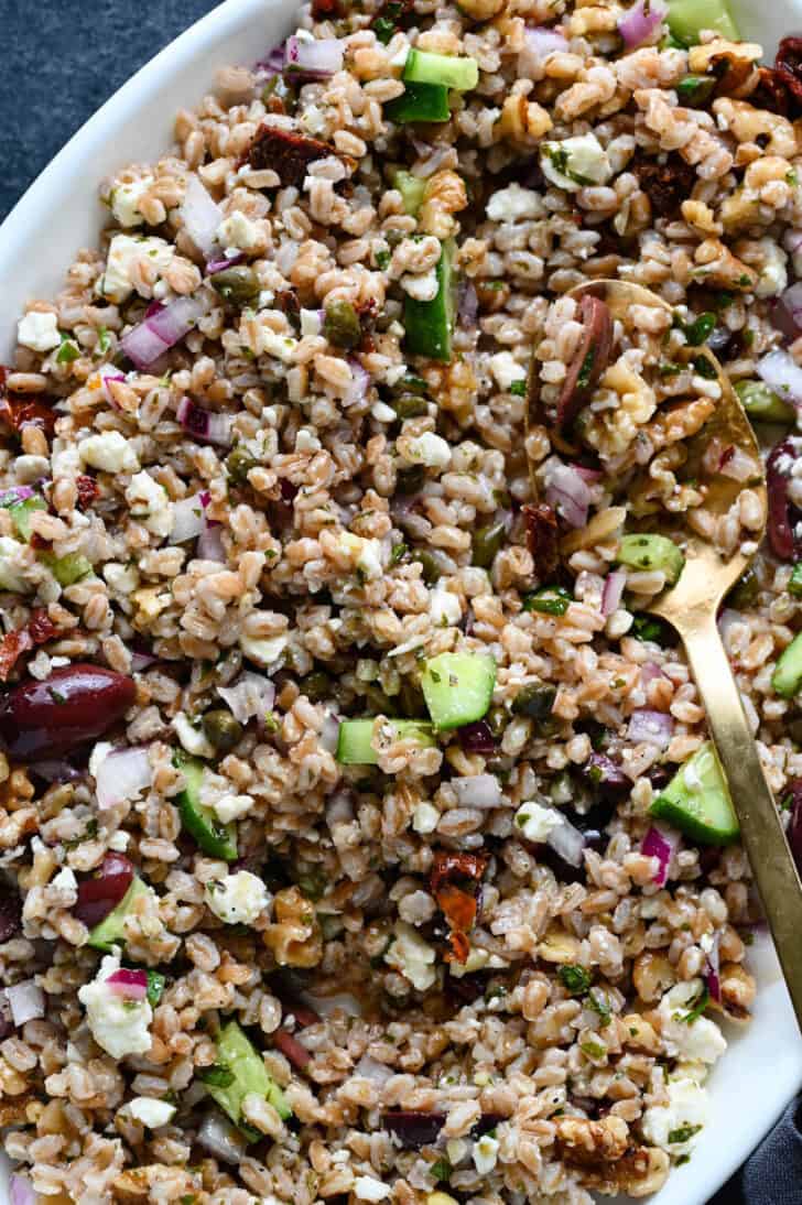 A grain and vegetable mixture on a white platter with a gold serving spoon digging in.