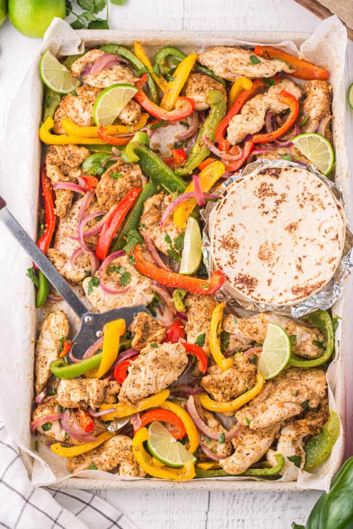 Sheet pan chicken fajitas with flour tortillas, and a metal spatula lifting some of the fajita mixture off the pan.