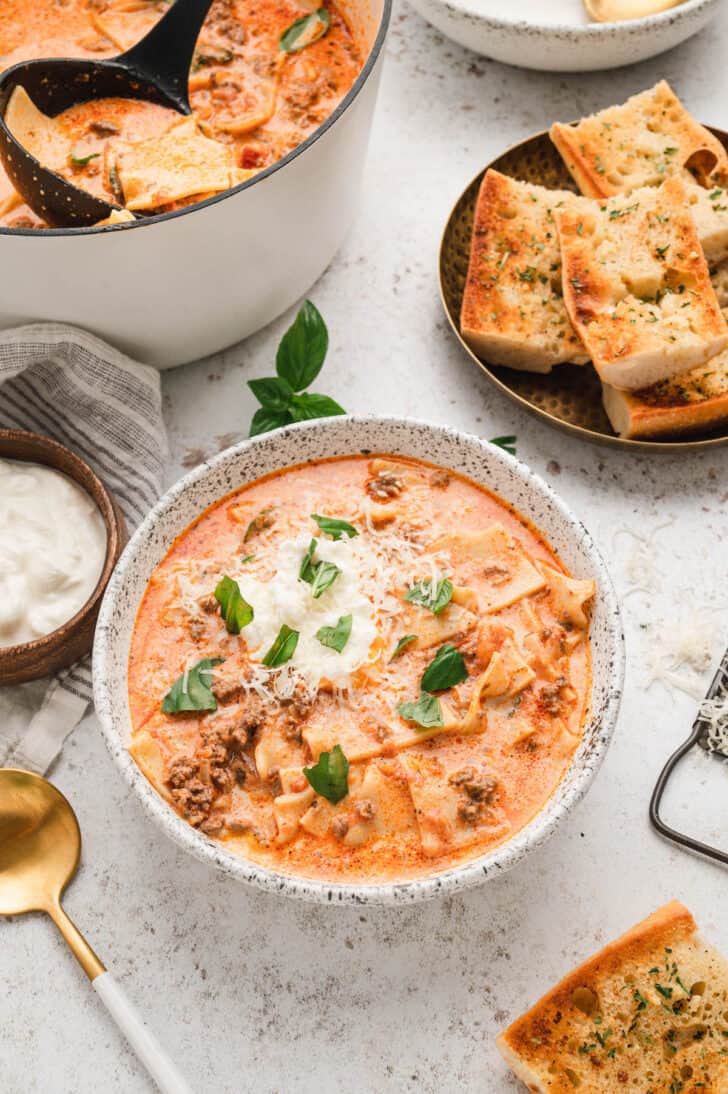 Lasagna Soup Recipe in a rustic bowl, topped with ricotta, Parmesan cheese and fresh basil, with garlic bread in the background.