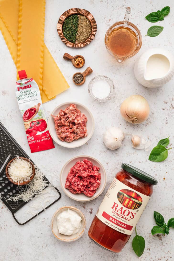 Ingredients laid out on a light surface, including pasta, ground meat, herbs, spices, onion, garlic, tomato paste, marinara sauce, cream and cheese.