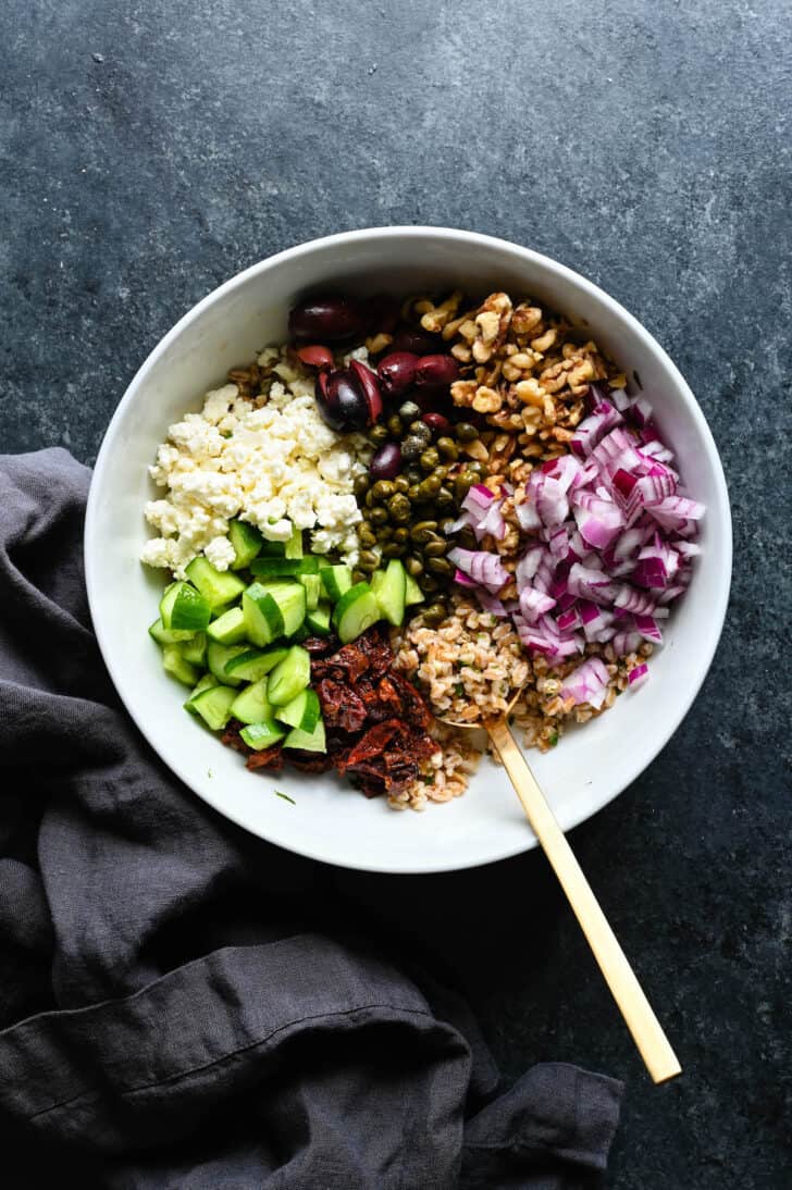 A farro salad recipe in a bowl but not yet stirred together, including cucumbers, sun-dried tomatoes, feta, capers, olives, walnuts and red onions.