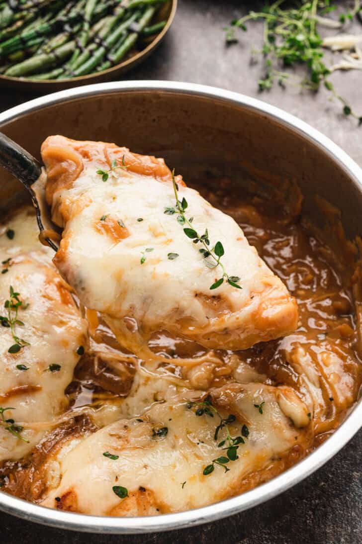 Chicken covered in white melted cheese being lifted out of a pan of caramelized onions.