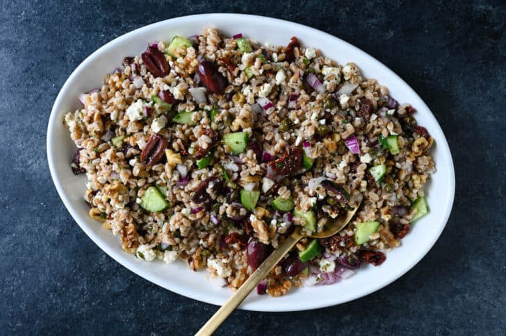 Mediterranean farro salad on a white platter with a gold serving spoon digging in.