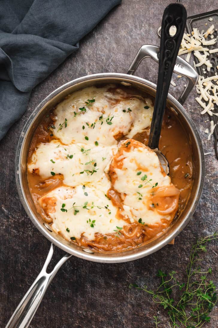 French Onion Baked Skillet in a stainless steel skillet with a black spoon lifting a piece of chicken out.
