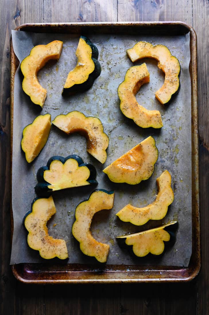A rimmed baking pan filled with acorn squash half moon pieces sprinkled with salt and pepper.