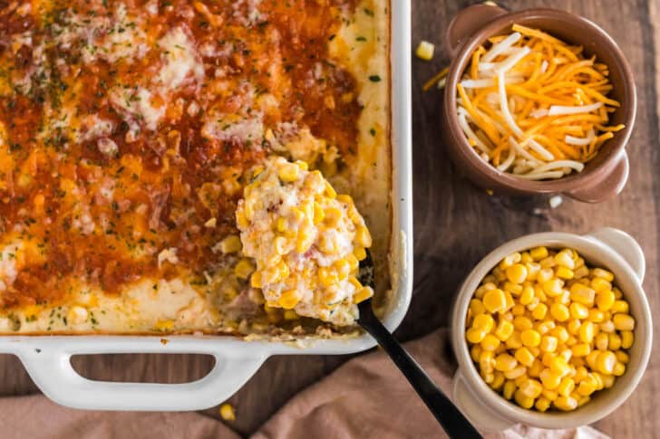 A white baking dish filled with a sweetcorn bake with cheese, with a black spoon lifting out a scoop.