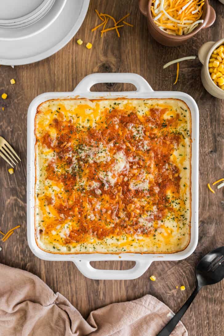 A cheesy corn casserole recipe in a square white baking dish on a wooden table.