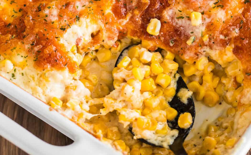 A white baking dish full of cheesy corn casserole with a black spoon taking a scoop out.