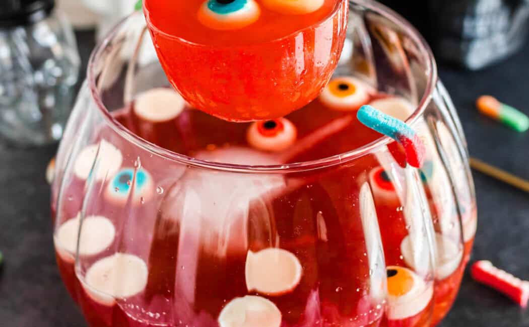 A glass bowl filled with red Halloween punch and gummy eyeballs, with a ladle lifting a serving out.