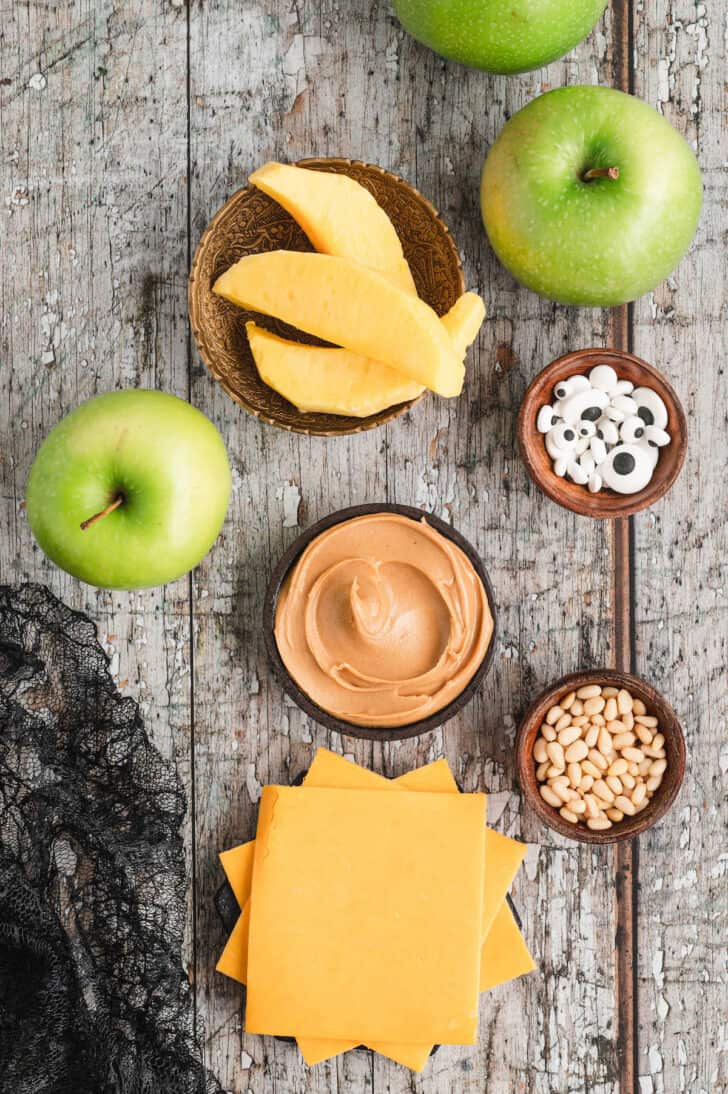 Ingredients on a rustic background, including green apples, peanut butter, cheese slices, sliced mango, pine nuts and candy eyeballs.