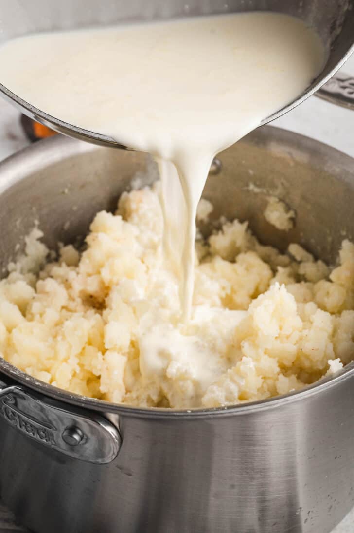 One pan pouring cream into another pan with crushed spuds in it.