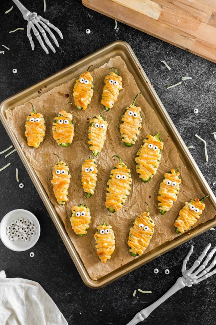 Jalapeno mummies with candy eyeballs on a parchment paper lined baking pan.