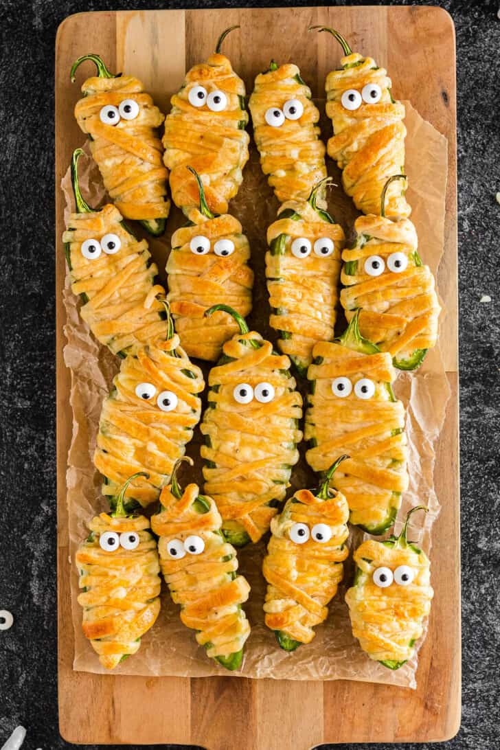 Jalapeno mummy poppers with candy eyeballs on a wooden cutting board.