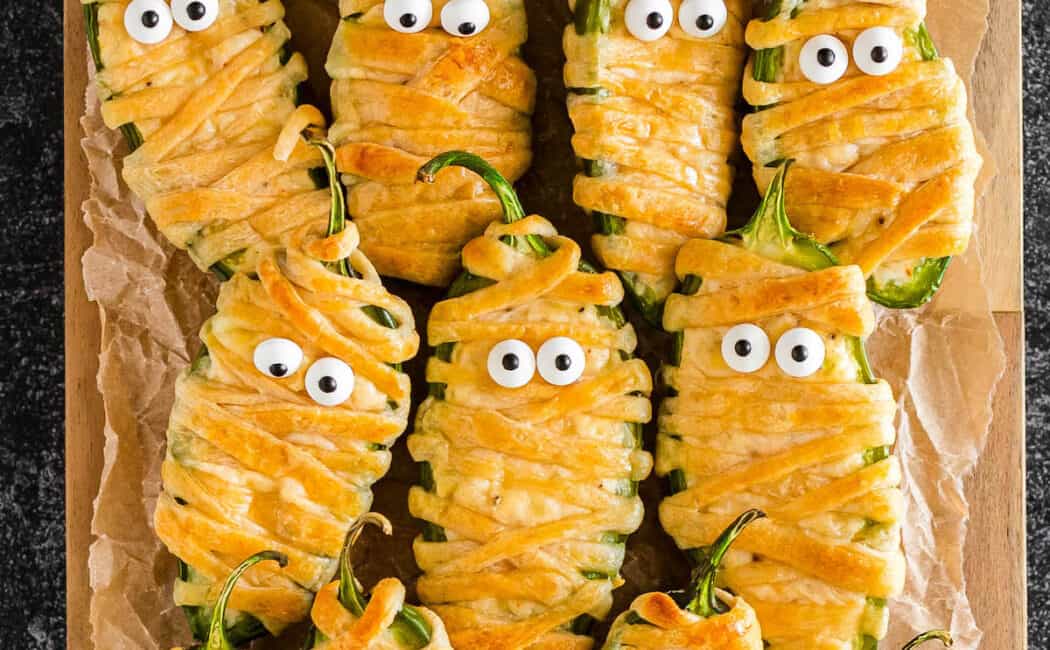 Mummy jalapeno poppers with candy eyeballs on a wooden cutting board.