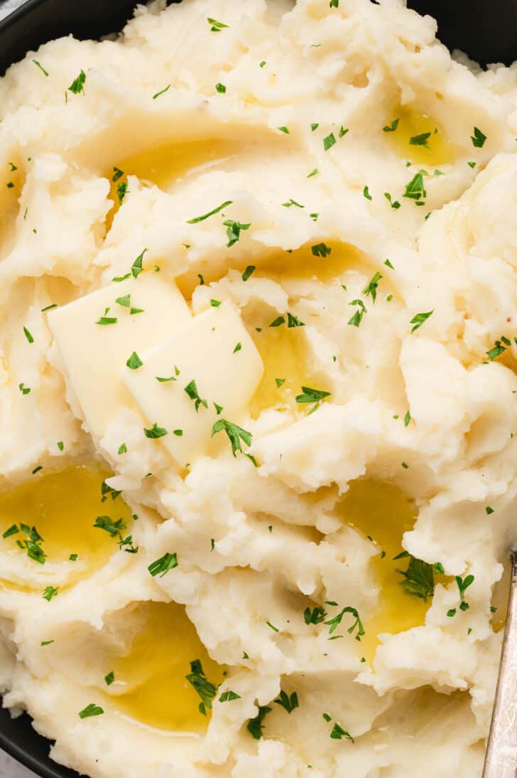 Homemade mashed potatoes in a black serving bowl garnished with pats of butter and chopped parsley.