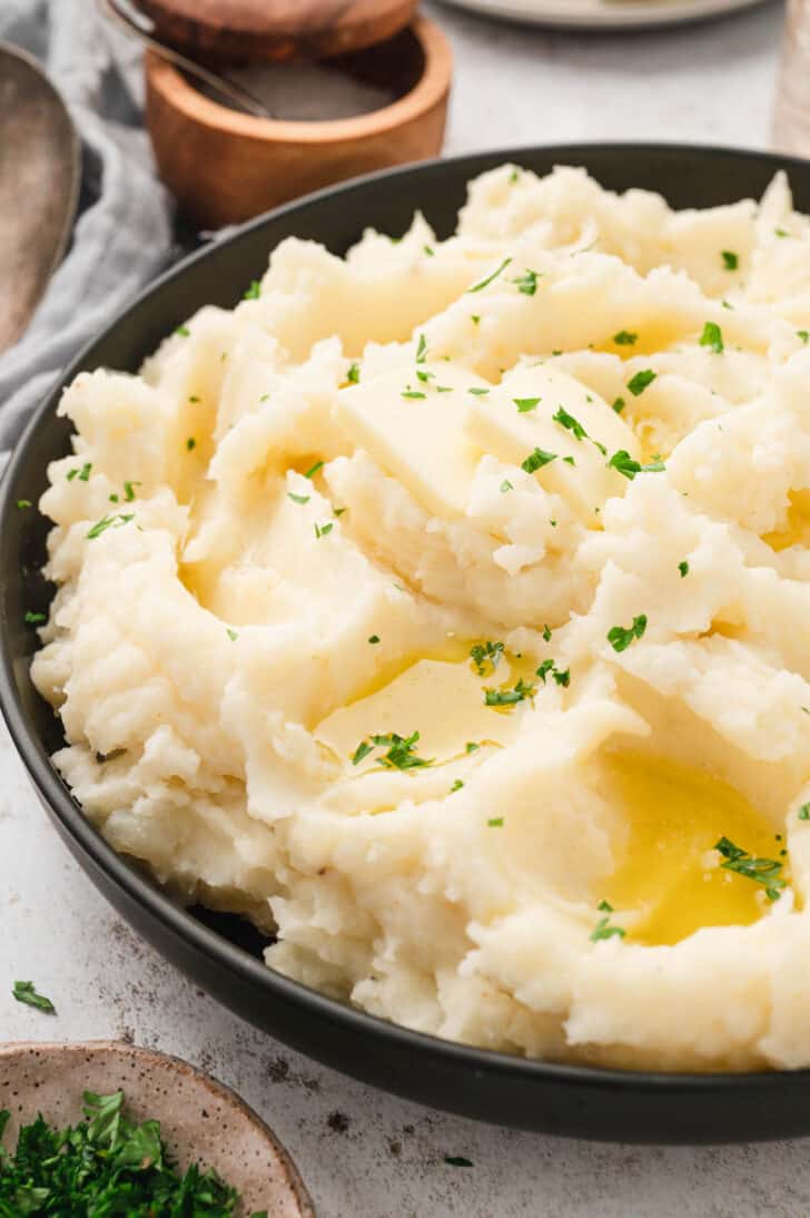 Mashed potatoes with heavy whipping cream in a black serving bowl garnished with pats of butter and chopped parsley.