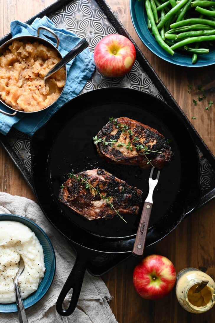 A cast iron skillet with pork chops with applesauce in a bowl alongside, as a meal scene with green beans and mashed potatoes.