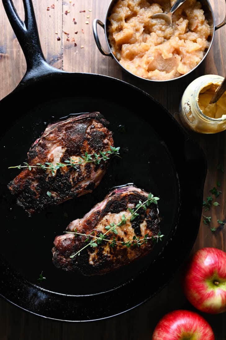 Two applesauce pork chops in a cast iron skillet, garnished with thyme.