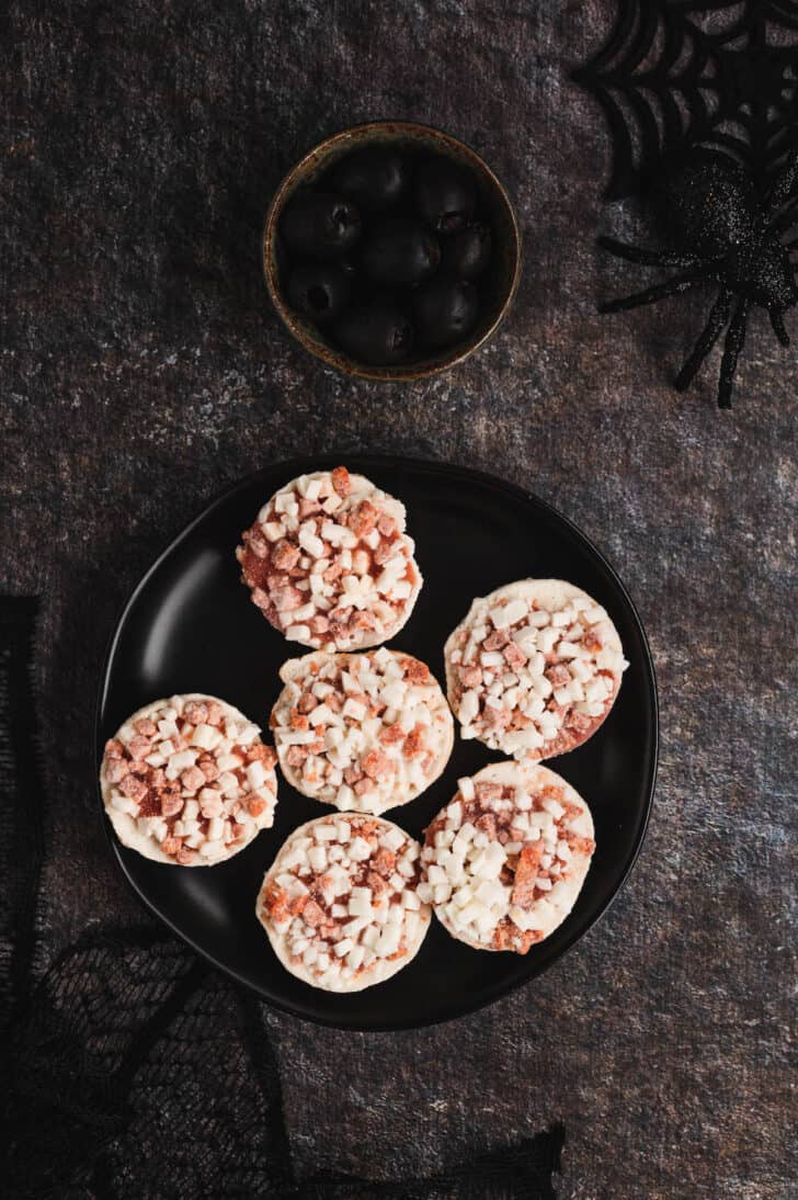 A plate of frozen mini pizzas and a bowl of black olives on a textured surface.